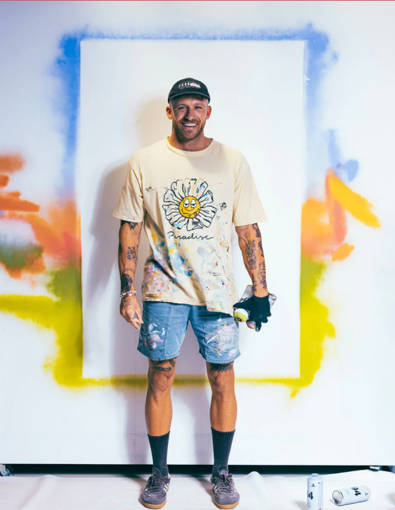 Man wearing a beige t-shirt with a sun design and denim shorts standing in front of a colorful abstract painting.