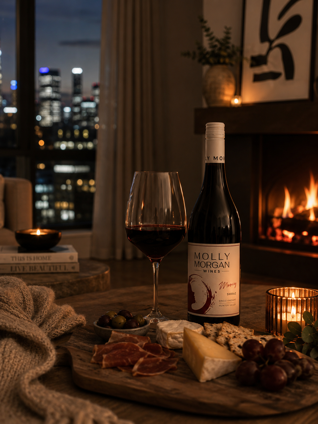 A bottle of Molly Morgan Winery Moroney Shiraz  wine and a filled wine glass on a rustic wooden table set in a cozy living room by a fireplace, with cheese, crackers, grapes, and a city skyline visible in the background.
